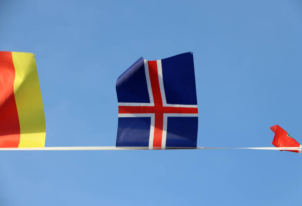 Mini fabric rail flag of Iceland in The Red Cross crosses over the white cross on the blue color hanging on the rope cloth between the flag of other countries and flick up, on blue sky background. - Photo, Image