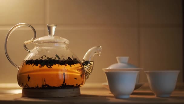 Tetera con hojas de té fresco en agua caliente y tazas pequeñas en la mesa de la cocina. Concepto. Primer plano de la tetera transparente de vidrio con la elaboración de té negro, concepto de ceremonia del té. - Metraje, vídeo