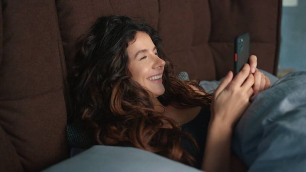 Boldog nő mobilt használ az ágyban. Vidám lány keres mobiltelefon képernyőn. - Fotó, kép