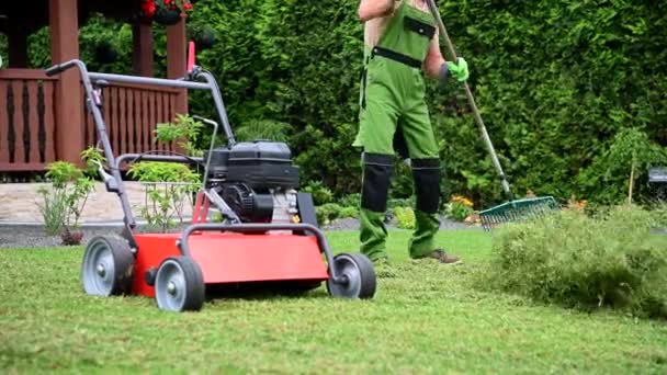 Miespuolinen työntekijä siivoamassa nurmikon leikkeitä leikkaamisen jälkeen asuinalueella. Puutarha huolto ruohonleikkuri ja rake.  - Materiaali, video