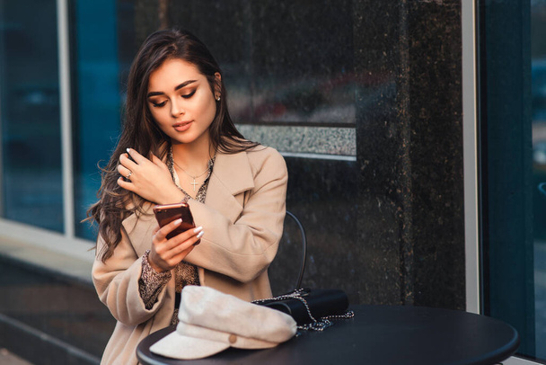 Women fashion. Woman with perfect makeup and hairstyle. Seductive girl at coffee shop outside. Technology and lifestyle. Fashionable girl with mobile phone sitting at cafe outdoors. City lifestyle. - Photo, Image