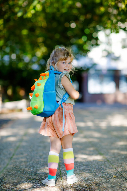 Słodka, mała, urocza dziewczynka na pierwszym dniu chodzenia do szkoły. Zdrowe, piękne dziecko spacerujące do przedszkola i przedszkola. Szczęśliwe dziecko z plecakiem na ulicy miasta, na zewnątrz. - Zdjęcie, obraz