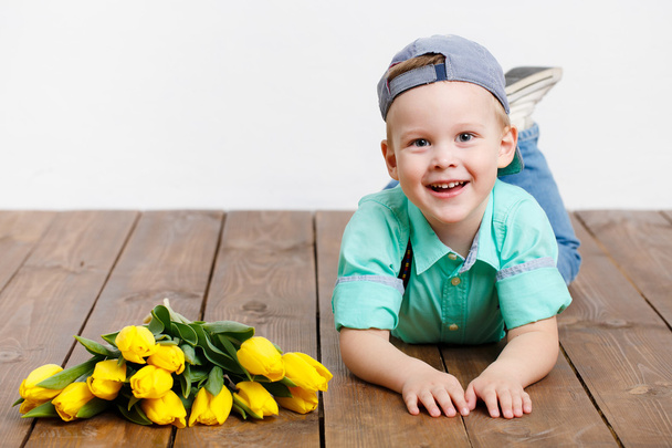 Smiling boy holding a bouquet of yellow tulips in hands sitting on wooden floor - Fotografie, Obrázek