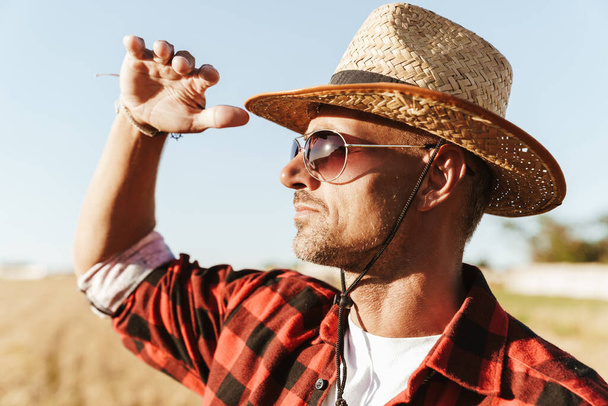 Image of unshaven adult man in straw hat and sunglasses looking aside while standing at cereal field - Valokuva, kuva