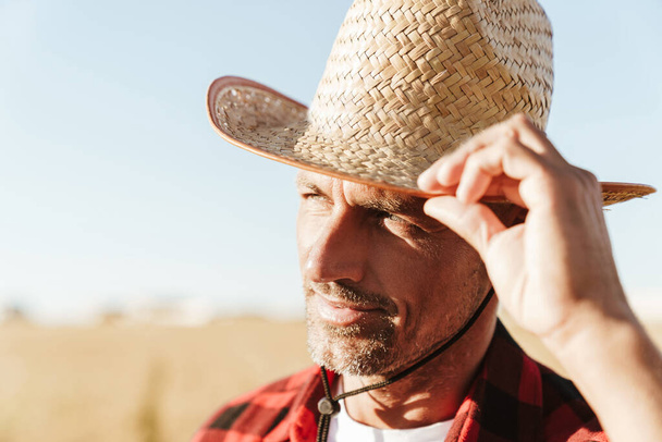 Image of unshaven adult man in straw hat looking aside while standing at cereal field - Foto, immagini