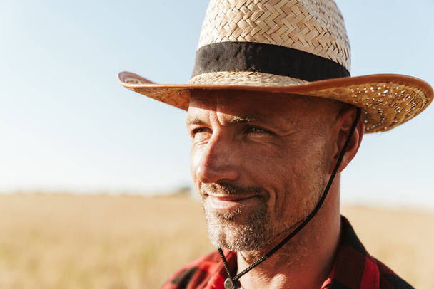 Image of pleased adult man in straw hat looking aside while standing at cereal field - Foto, immagini