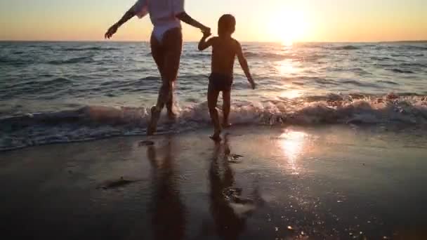 Mujer joven y su hijo vienen al agua en la puesta de sol en la playa de arena, vacaciones en la isla - Imágenes, Vídeo