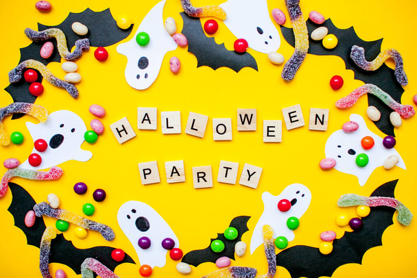 inscription from wooden blocks Halloween party and frame made of paper homemade bats and paper ghosts and multicolored candies and worms from gummy on a bright yellow background - Photo, image