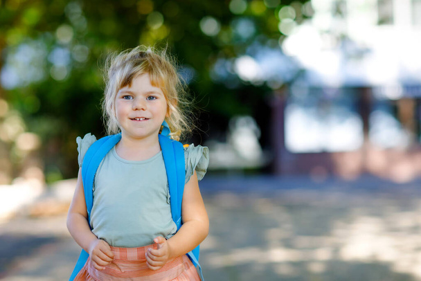 Aranyos kis aranyos kislányka az első napján, aki játszóiskolába jár. Egészséges gyönyörű baba sétál óvodába és óvodába. Boldog gyerek hátizsákkal a város utcáján, szabadban. - Fotó, kép
