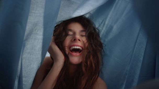 Χαρούμενο κορίτσι που ποζάρει κάμερα κάτω από ντυμένο ύφασμα. Γυναίκα που γελάει κάτω από σεντόνι. - Φωτογραφία, εικόνα