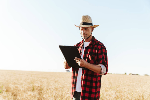 Image of pleased adult man in straw hat working with papers while standing at cereal field - Foto, immagini