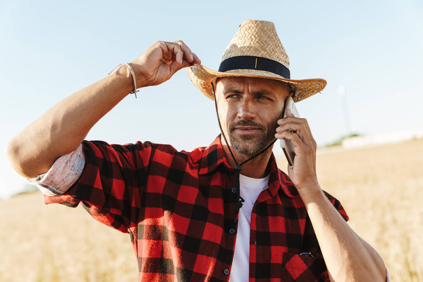 Image of unshaven adult man in straw hat talking on cellphone while standing at cereal field - Foto, Imagen