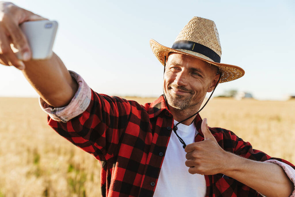 Image of pleased adult man in straw hat taking selfie on cellphone while showing thumb up at cereal field - Фото, зображення