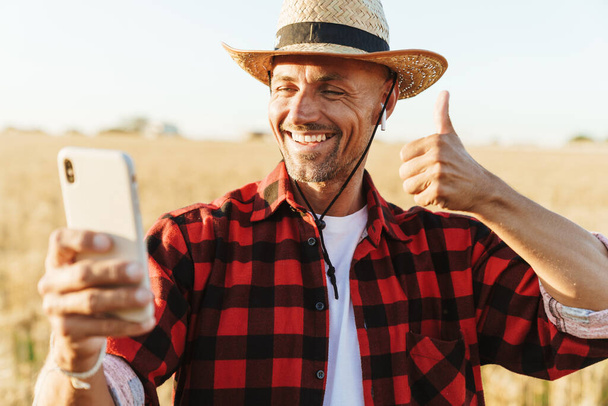 Image of smiling adult man in earphones showing thumb up and taking selfie on cellphone at cereal field - Fotografie, Obrázek