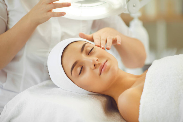 Smiling young womans face under light getting and enjoying facial massage from cosmetologist - Foto, Imagem