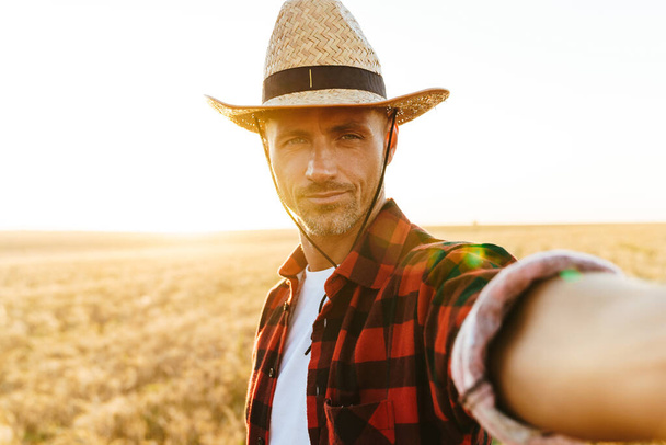 Image d'un bel homme adulte en chapeau de paille prenant des photos selfie tout en se tenant debout au champ de céréales - Photo, image