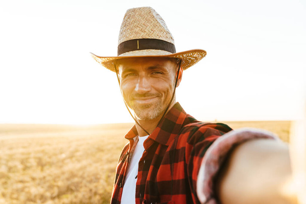 Image of pleased adult man in straw hat taking selfie photo while standing at cereal field - Φωτογραφία, εικόνα