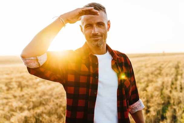 Изображение довольного взрослого мужчины, улыбающегося и смотрящего в камеру, стоя на зерновом поле - Фото, изображение
