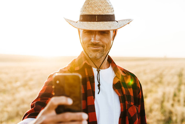 Image of pleased adult man in straw hat taking selfie on mobile phone at cereal field - Photo, Image