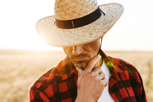 Image of unshaven adult man in straw hat looking downward while standing at cereal field - Foto, Imagen