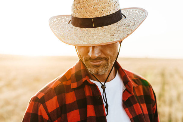 Image of unshaven adult man in straw hat looking downward while standing at cereal field - 写真・画像