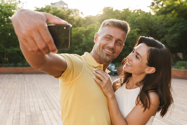 Portrait of joyful adult couple man and woman taking selfie photo on cellphone and hugging while walking in summer park - Foto, Bild