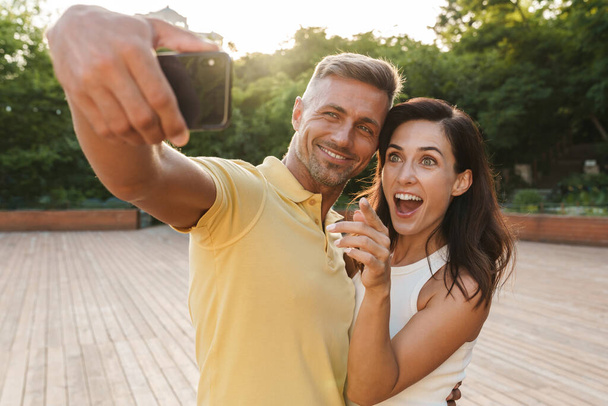 Portrait of excited adult couple man and woman taking selfie photo on cellphone and pointing finger while walking in summer park - Fotó, kép