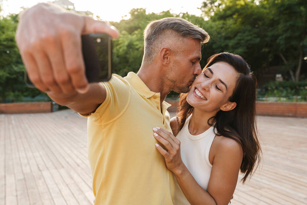 Portrait of pleased adult couple man and woman taking selfie photo on cellphone and kissing while walking in summer park - Fotó, kép