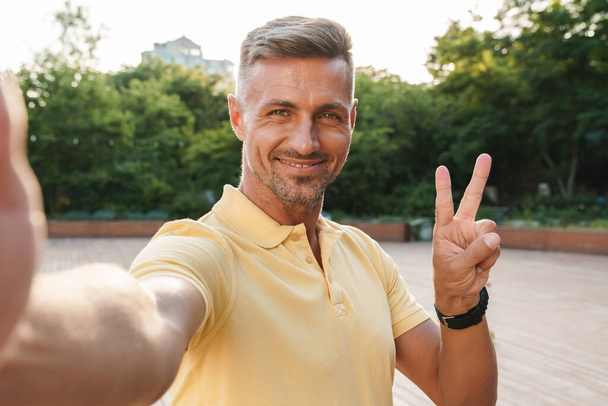 Image closeup of smiling middle-aged man wearing yellow t-shirt taking selfie photo and gesturing peace sign while walking in summer park - Foto, immagini