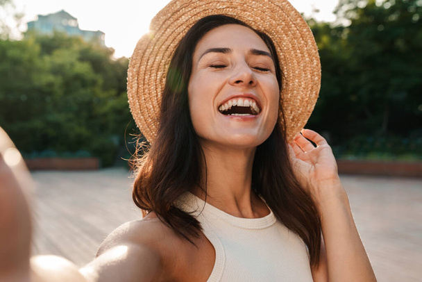 Image of nice middle-aged woman wearing straw hat taking selfie photo and laughing while walking in summer park - 写真・画像