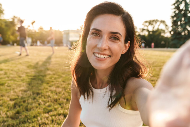 Image of smiling middle-aged woman taking selfie photo and looking at camera while sitting on grass in summer park - Photo, Image