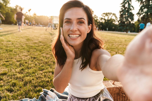Image of positive middle-aged woman taking selfie photo and looking at camera while sitting on grass in summer park - Foto, afbeelding
