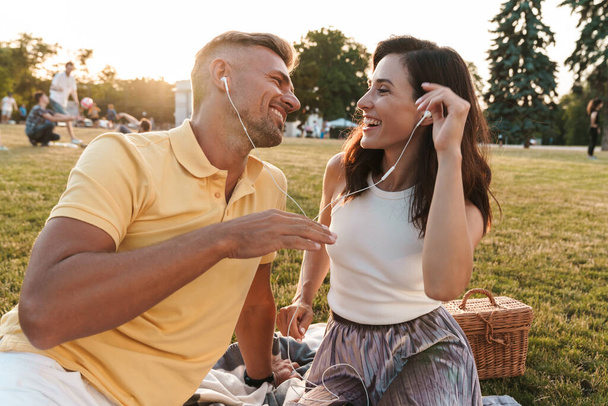 Portrait of beautiful middle-aged couple man and woman laughing and listening to music together with earphones while sitting in summer park - Fotografie, Obrázek