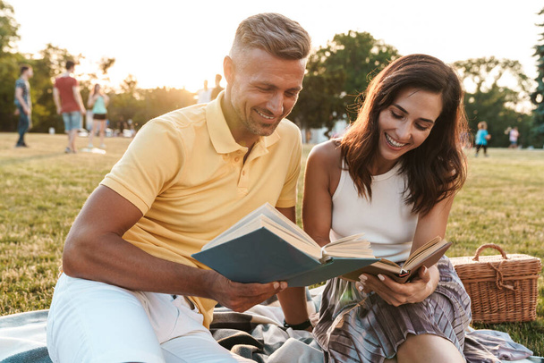 Portrait of happy middle-aged couple man and woman smiling and reading books together during picnic in summer park - Photo, image