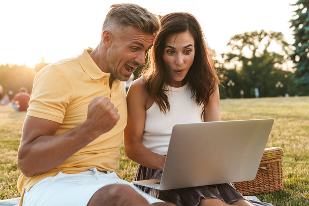 Portrait of surprised middle-aged couple man and woman screaming and celebrating success while using laptop computer in summer park - Foto, Bild