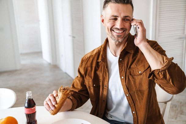 Image of grey-haired joyful man smiling and talking smartphone while have lunch in white kitchen - Foto, Imagem