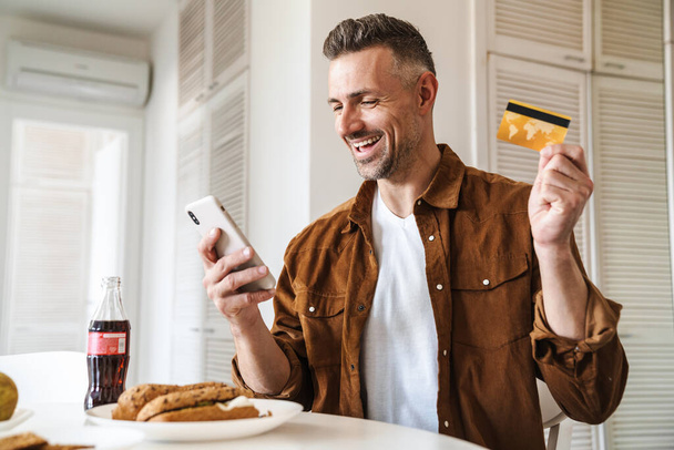 Image of handsome joyful man holding credit card and using smartphone while have lunch in white kitchen - Photo, image