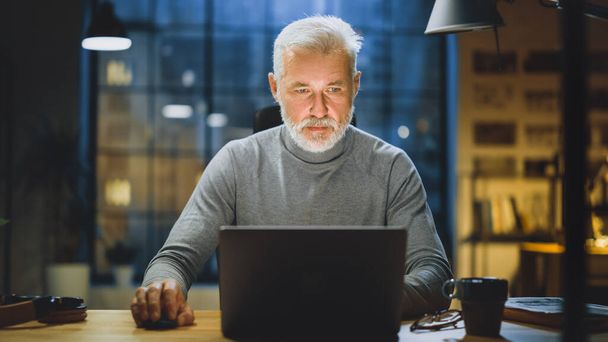 Portrait of the Handsome and Successful Middle Aged Bearded Businessman Working at His Desk Using Laptop Computer. Working from Cozy Home Office Studio with Window View of the City at Night - 写真・画像