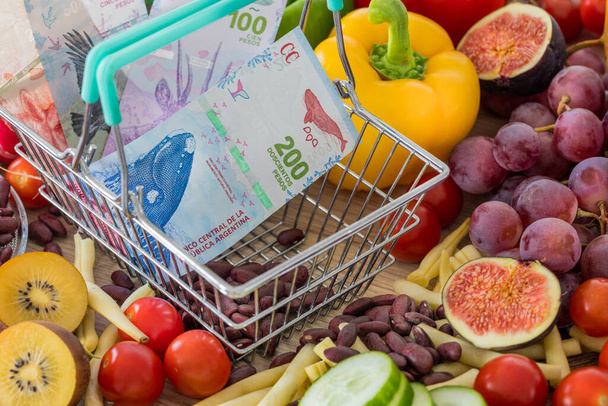 Cesta de compras con dinero argentino, alrededor de productos alimenticios, verduras y frutas. El concepto de inflación, aumento de precios y alimentos más caros - Foto, Imagen