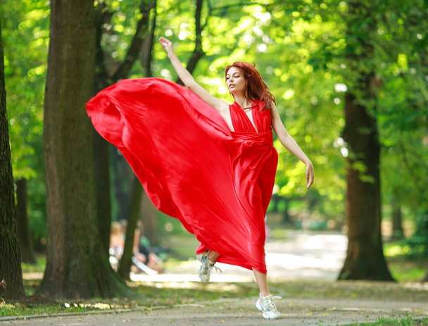 Young fashionable woman in long red luxurious dress walks in a summer park - Φωτογραφία, εικόνα