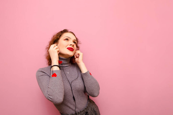 Happy girl with curly hair listens to music in headphones with a smile on her face, looks up at copy space, isolated on pink background, wears gray sweater. - Photo, image
