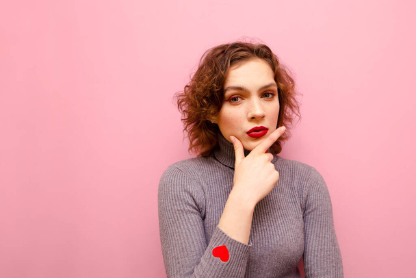 Closeup portrait of pensive girl in gray sweater on pink background, looking into camera and thinking. Curly girl with a pensive face isolated on a pink background,looks in camera with a serious look - Foto, imagen