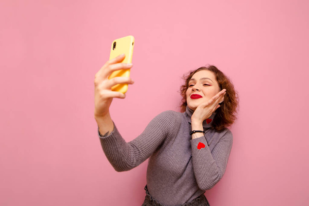 グレーのタートルネックに巻き毛の可愛い女の子がピンクを背景に自撮りをしています。スマートフォンのカメラで写真を撮ることに積極的な女性。スペースのコピー - 写真・画像