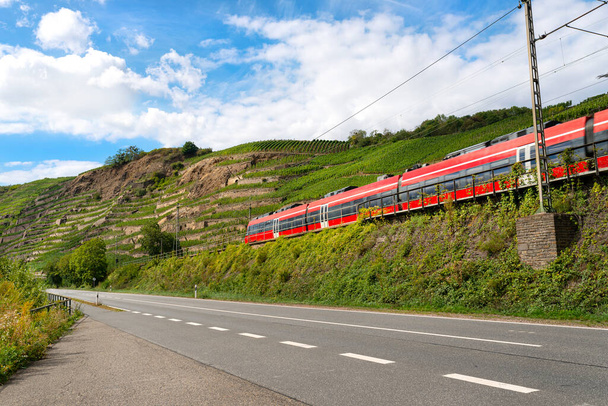 Un tren rojo de pasajeros que conduce junto a una autopista vacía junto a las colinas de un viñedo en Alemania. - Foto, imagen
