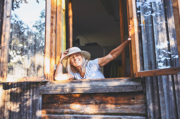 Šťastný úsměv emocionální starší žena baví pózování otevřeným oknem v rustikální staré dřevěné vesnice domu v slamáku klobouk. Koncept důchodců. Karanténa v domě na venkově - Fotografie, Obrázek