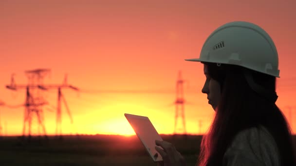 Een vrouwelijke power engineer in witte helm inspecteert de stroomleiding met behulp van gegevens van elektrische sensoren op een tablet. Hoogspanningsleidingen bij zonsondergang. Distributie en levering van elektriciteit. schone energie - Video