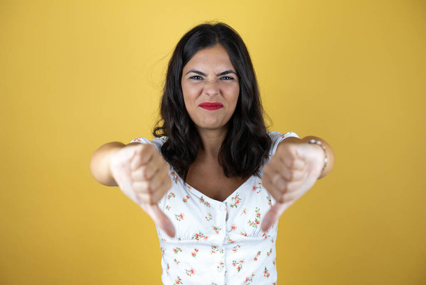Красивая женщина на желтом фоне с сердитым лицом, негативный знак показывает неприязнь с опущенными пальцами - Фото, изображение