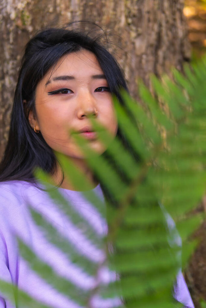 Portrait of a young Asian brunette in a park next to a tree and a fern leaf near her face. Lifestyle, portrait Asian girl - Fotoğraf, Görsel