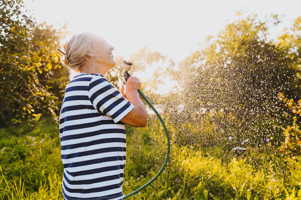 Happy elderly senior woman having fun watering plants with hose in summer garden. Drops of water in backlight. Farming, gardening, agriculture, old age people. Growing organic vegetables on farm.  - Photo, image