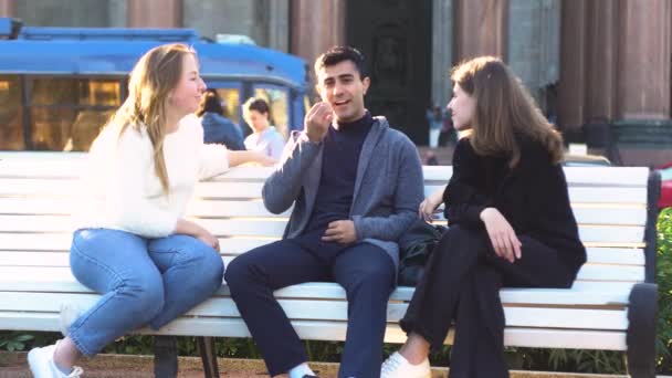 Dwie młode dziewczyny i mężczyzna przyjaciele siedzą na ławce w letnim parku i rozmawiają szczęśliwie dobrze się bawiąc. Media. Portret dwóch uśmiechniętych kobiet i mężczyzny spędzających razem czas i rozmawiających. - Materiał filmowy, wideo
